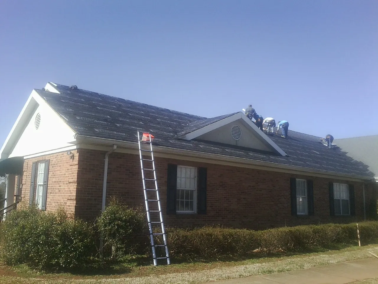 Crew tearing shingles off a brick ranch roof with a ladder set — Metal Roof Repair work by our crew in Big Rapids